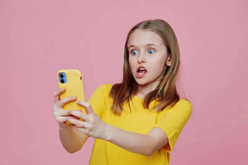 Surprised girl using smartphone, wearing yellow t shirt, pink background, expressing shock and excitement, modern concept of digital communication