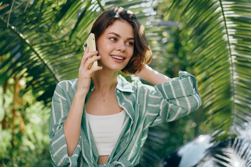 Young woman outdoors talking on a smartphone, wearing a striped green shirt and white crop top, surrounded by tropical plants.