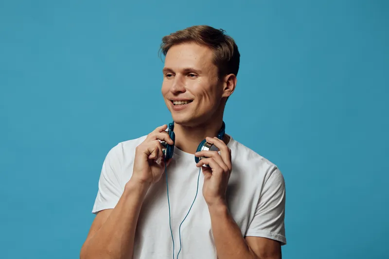 Smiling young man with headphones holding earphones in hand, wearing casual white t-shirt, isolated on blue background, studio portrait, cheerful mood, lifestyle concept, modern technology