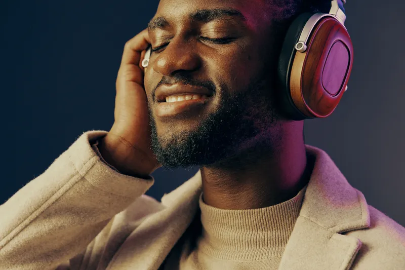 Joyful Black man enjoying music with headphones on a dark background, conveying relaxation and happiness while dressed in a stylish beige sweater coat