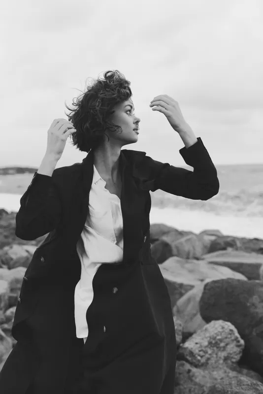 Woman in blazer and white shirt on a rocky coastline, wind in her hair, monochrome fashion portrait capturing movement, confidence, and sea breeze toned.