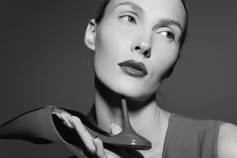 Portrait of a fair skinned Caucasian woman holding a high heel shoe near her face. Studio shot in black and white with dramatic lighting and a contemplative expression.