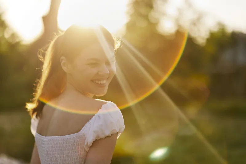 Young woman with a joyful smile in a sunlit field, wearing a white dress, showcasing happiness and natural beauty in a serene outdoor setting
