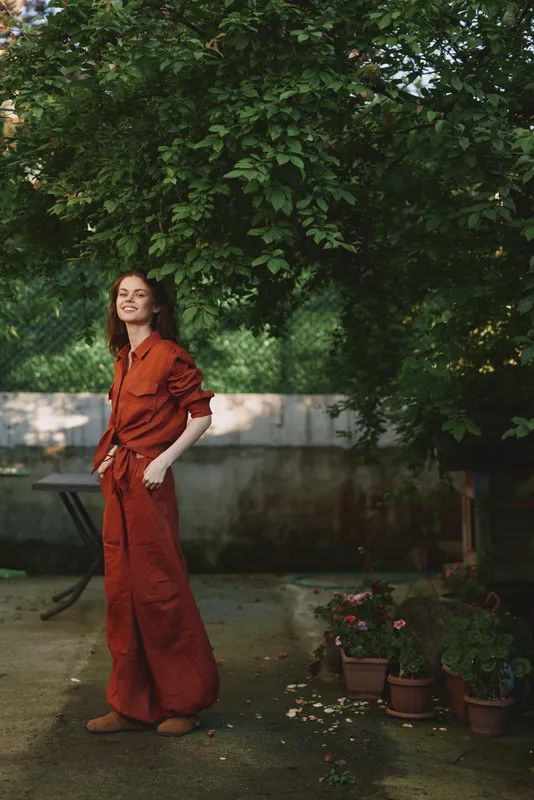 Smiling woman wearing an orange jumpsuit stands outdoors in a garden, sunlight filters through leaves, highlighting a relaxed lifestyle, casual fashion and natural confidence, perfectly framed by