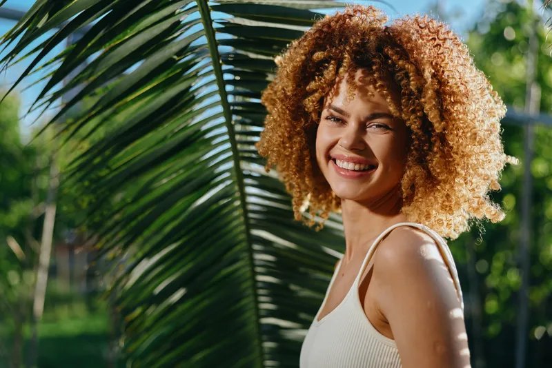 Smiling woman with curly hair enjoys sunny day outdoors near green palm leaf, wearing white tank top with natural light and cheerful summer mood.