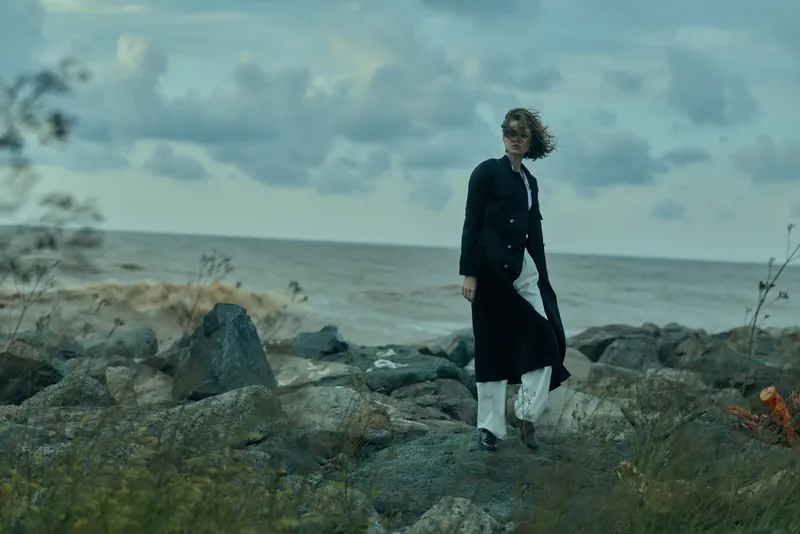 Windy coastal scene with a woman walking on a rocky shore, wearing a long coat, as the sea and cloudy skies create a moody atmosphere.