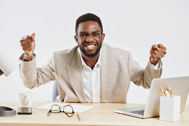 Smiling happy man with African descent in business casual attire celebrating success or achievement at work, sitting at desk with laptop, glasses, notepad, and office supplies, white background studio