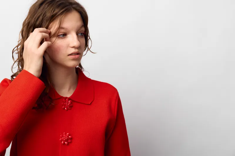 Young woman in red sweater looks away thoughtfully, touching her hair. Isolated on white background. Concept of contemplation, casual fashion, and natural beauty portrait.