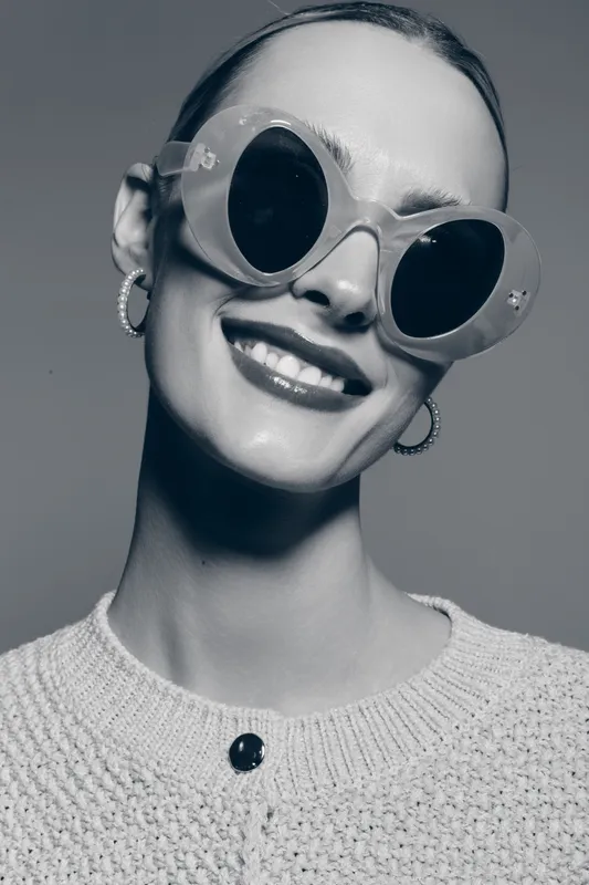 Smiling caucasian woman with light skin wearing large round sunglasses and hoop earrings. She wears a knitted sweater and poses confidently on a plain grey background with natural lighting.