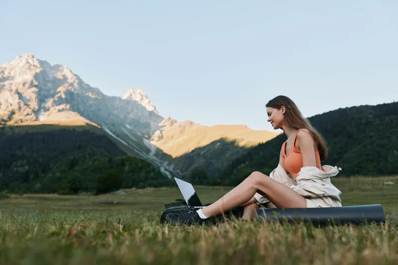 A focused young woman sits on a yoga mat in a scenic meadow with a laptop, enjoying remote work in nature. The sunny mountain backdrop conveys freedom and productivity.