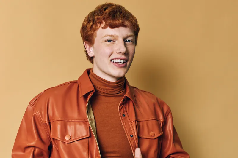 Portrait of smiling young man with red hair wearing brown leather jacket and turtleneck against beige background in studio for fashion and lifestyle.