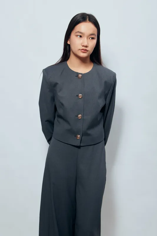 Young woman in dark gray suit standing with hands behind back on plain background. Asian female model with long hair looking sideways in minimalist studio portrait fashion style.
