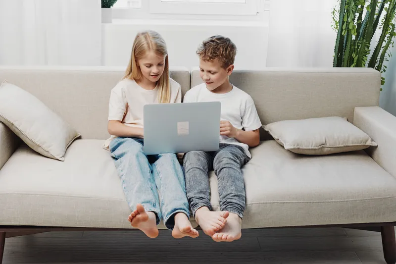 Two children sitting barefoot on a sofa using a laptop together in a bright living room. Concept of learning, technology, childhood, and casual home environment.