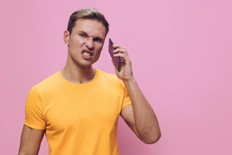 frustrated angry young caucasian man hold smartphone wear yellow t-shirt isolated pink background digital detox concept communication technology