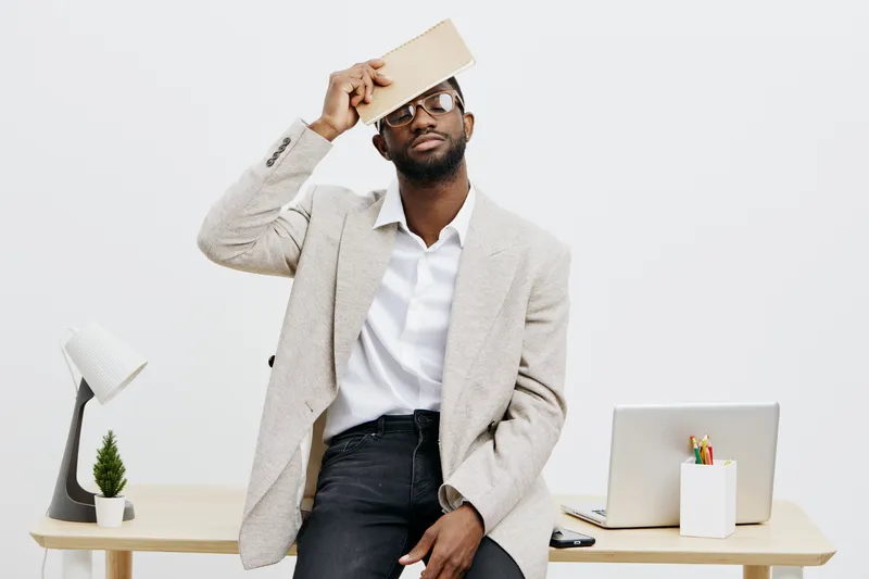 Confident young man of African descent with beard, wearing glasses and smart casual outfit, holds book over his head while standing in modern office space with white background, laptop and desk access