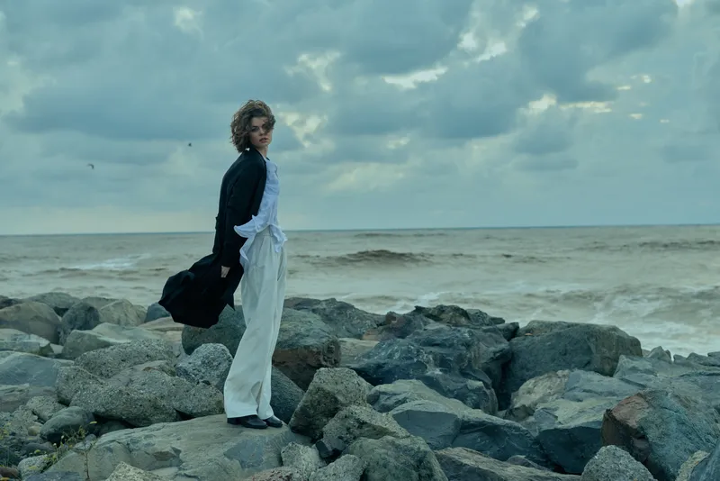 Woman wearing light overalls and dark jacket stands on rocky shoreline by the ocean, wind in her hair, moody sky over a coastal landscape