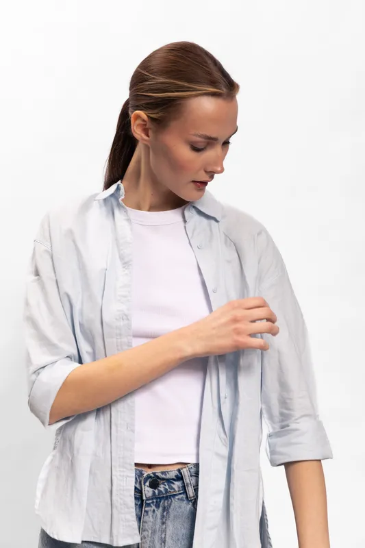 Young woman with brown hair in casual white shirt and light purple top looks down while adjusting sleeve. Minimalist studio portrait with white background and natural expression.