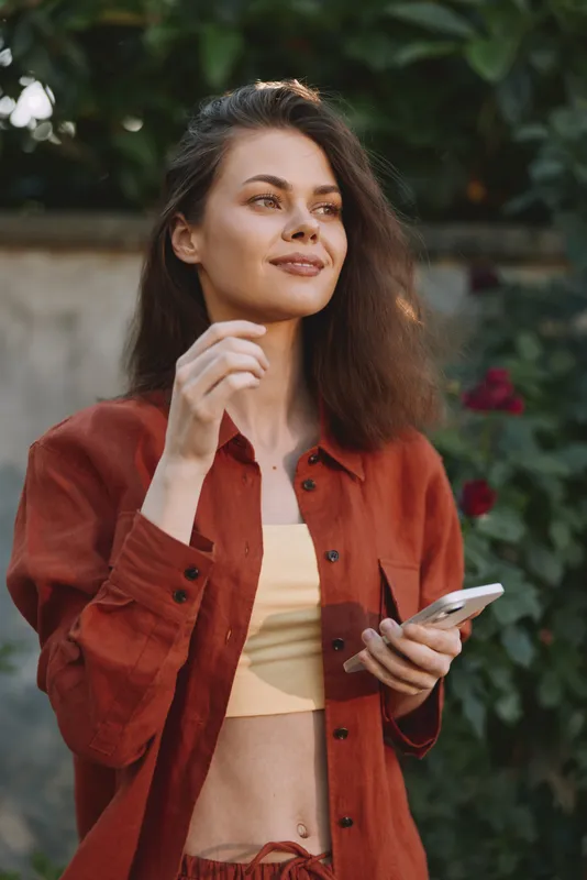 Young woman with wavy brown hair stands in a sunny garden, wearing a rust jacket over a pale yellow crop top, gently touching her hair while holding a smartphone. Her relaxed pose and soft smile