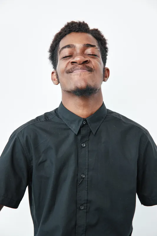 Young black man with dark skin wearing black shirt posing with confident smile and closed eyes on white background. Portrait of African ethnicity male with short hair and facial hair.