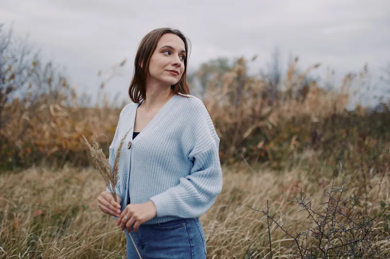 Candid portrait of a young woman standing in a windswept meadow, wearing a cozy cardigan and holding dried grasses, her sincere expression and relaxed pose conveying authentic, natural presence.
