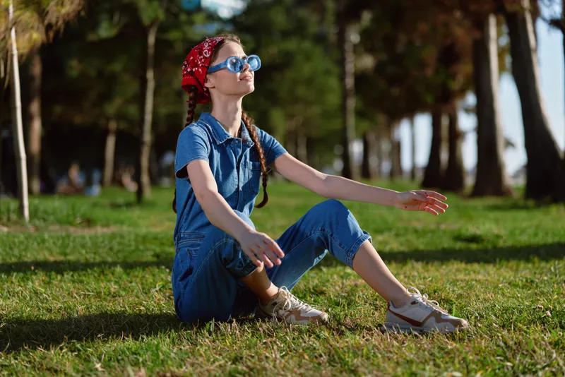 Lifestyle woman in modern boho-western denim street style sits relaxed on green grass in warm sunlight, wearing sunglasses and a red bandana, film color tone.