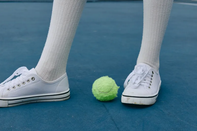 Young athlete wearing white sneakers and knee high socks standing on a blue tennis court with a bright green tennis ball, capturing the essence of sports and determination