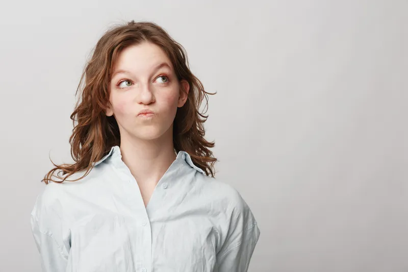 Young woman with curly hair in white shirt looks sideways with thoughtful expression on light background. Concept for doubt, decision making, and contemplation.