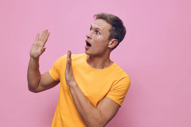 surprised man in yellow t-shirt looking away showing stop hands isolated on pink background digital detox concept emotion expression