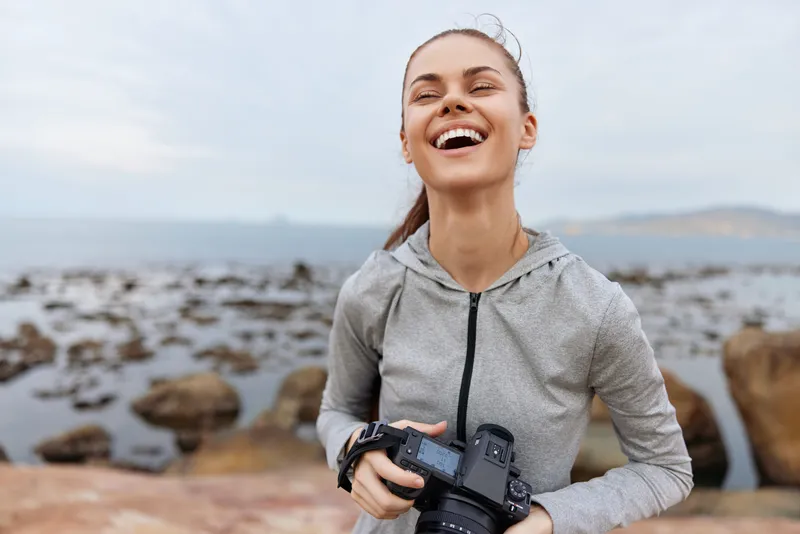 Young woman laughing joyfully while holding a camera on a rocky beach, wearing a grey sports jacket, capturing memories in nature s beauty