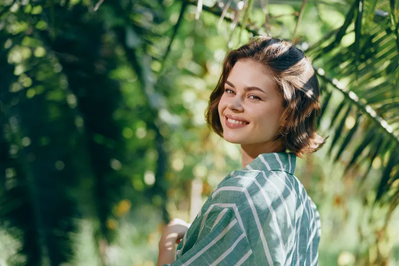 Cheerful woman smiles in natural light amid lush greenery, wearing a striped shawl, relaxed pose, bright mood.