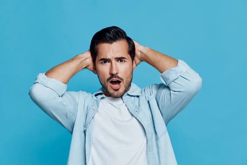 Surprised young man with dark hair express shock and excitement, wearing casual white t-shirt and denim jacket, against a vibrant blue background. Emotive lifestyle concept.