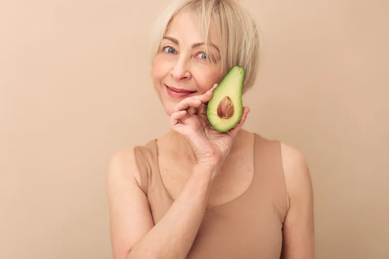Smiling mature woman with short blonde hair holds half of fresh avocado near face on beige background. Concept of healthy eating, natural skincare, wellness, and organic lifestyle for older adults.