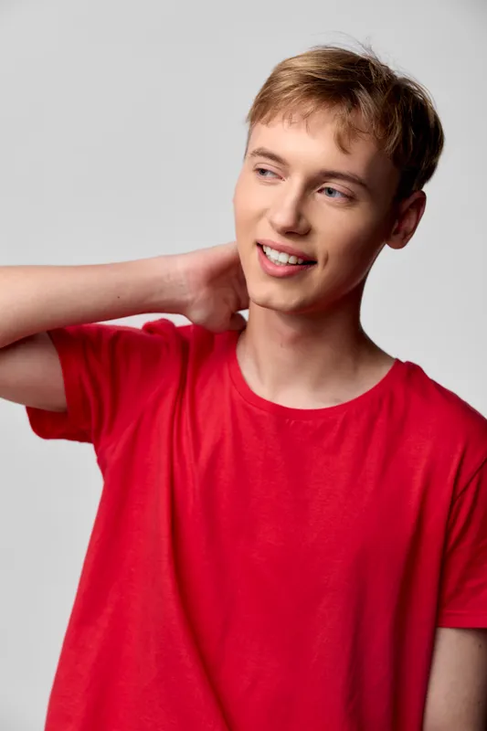 Male model wearing a bright red t shirt stands in a studio with a casual pose, hand at the neck and a warm smile, creating an approachable lifestyle fashion shot with a simple, clean background