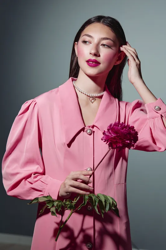 Young woman with bright lipstick wearing a pink creative fashion outfit holding a dark flower, posing with hand near hair. Beauty concept with bold makeup on grey background.