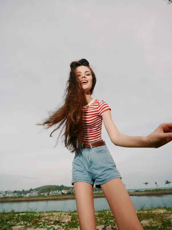 Energetic young woman wearing a red striped tee and high waisted denim shorts smiles as wind lifts her hair, outdoors by a calm lake under a bright sky.