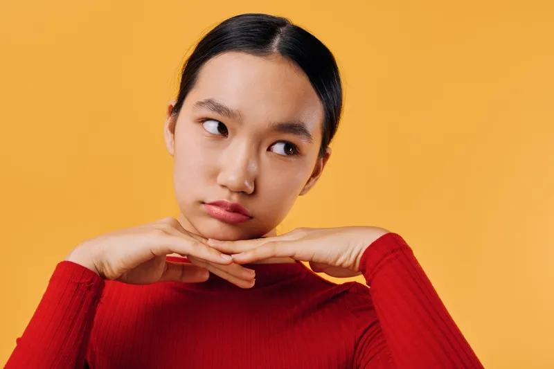 Young woman in red sweater with thoughtful expression posing with hands under chin on orange background. Portrait of pensive female with natural makeup and dark hair tied back.