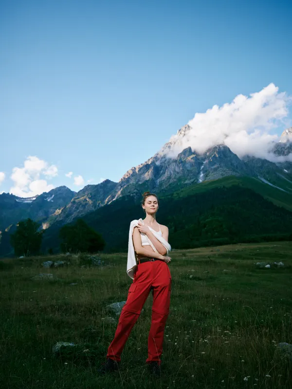 Mountain landscape, outdoor fashion on a model in a grassy field with red pants and white top, dramatic sky and distant peaks, nature and style in color