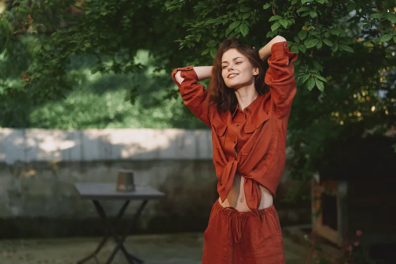 Playful woman outdoors, relaxed lifestyle moment, smiling with arms raised, sunlit scene, natural breeze, vibrant orange outfit, lush greenery backdrop, candid joyful vibe and natural charm