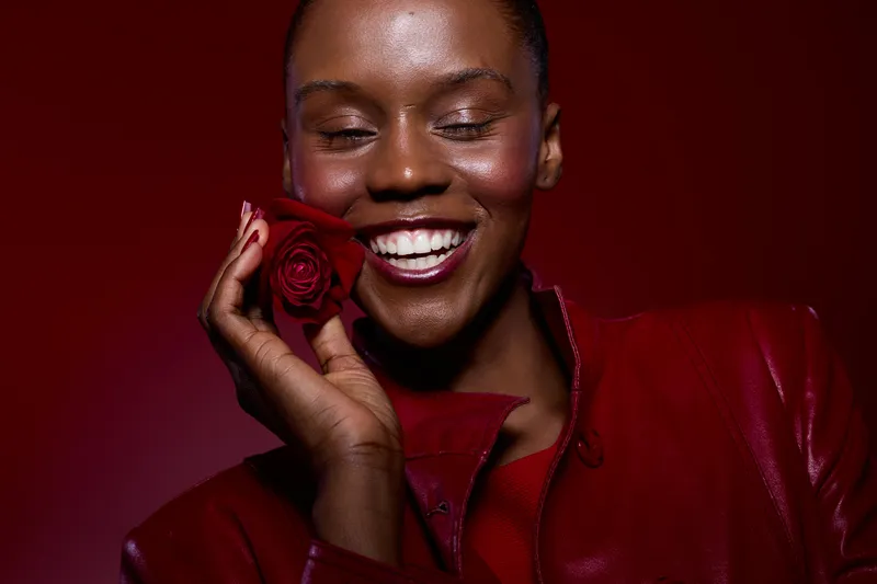 Smiling dark skinned African woman in red leather jacket holds red rose near face with closed eyes. Studio portrait on red background with natural makeup and joyful expression concept.