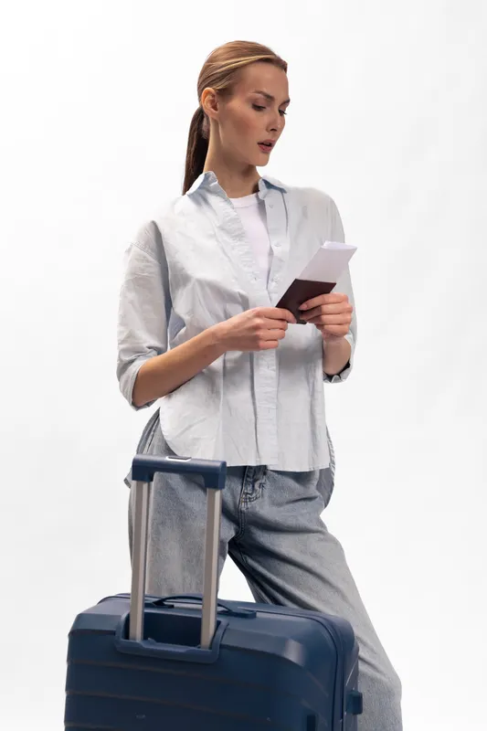 Young woman in casual clothes standing with blue suitcase reading passport and tickets. Travel concept with relaxed female traveler preparing for a journey on white background.