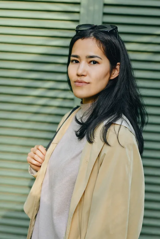 Woman wearing a beige coat and light sweater stands outdoors beside striped siding, calm expression, fashion casual lifestyle photography emphasizing timeless elegance and modern outerwear