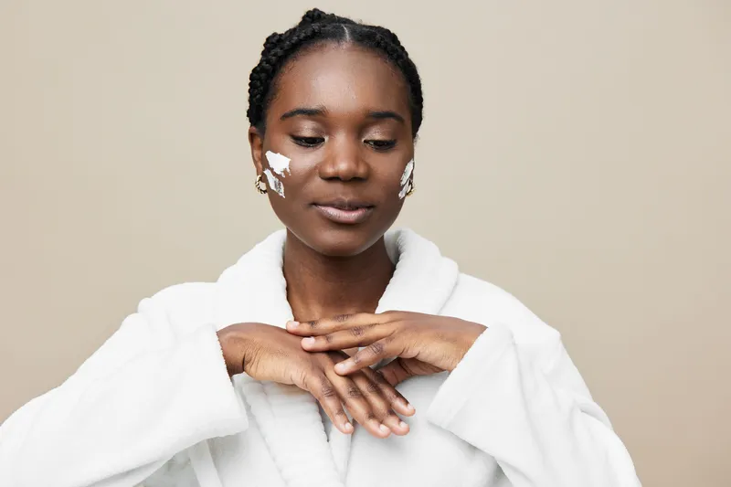 A serene Black woman applying skincare in a white robe, showcasing healthy skin and a calming atmosphere for wellness and beauty concepts