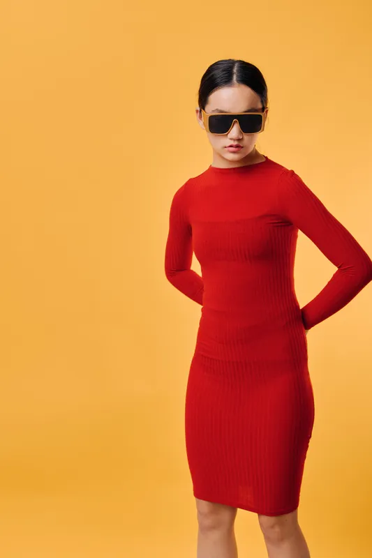 Woman wearing red dress and large sunglasses stands against orange background, posing with hands behind back. Fashion model, confident expression, studio shot for style and elegance.