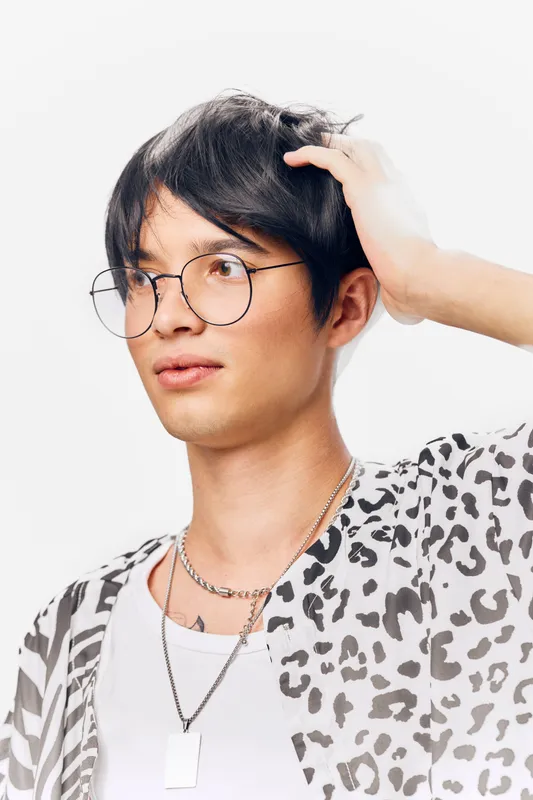 young man with black hair wearing glasses and leopard print shirt thoughtfully touching his head against white background in studio