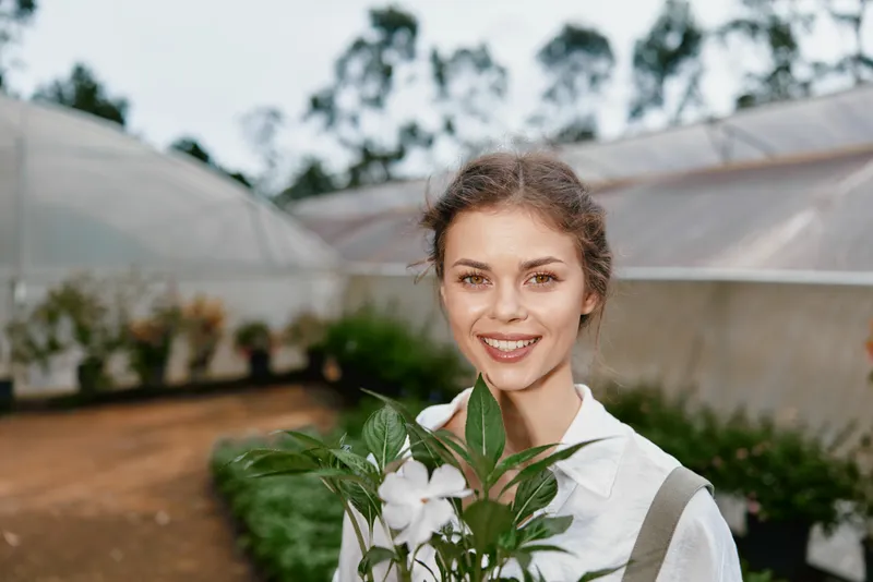 Young woman smiling in a greenhouse holding a plant, depicting joy and connection to nature, ideal for themes of gardening and sustainability