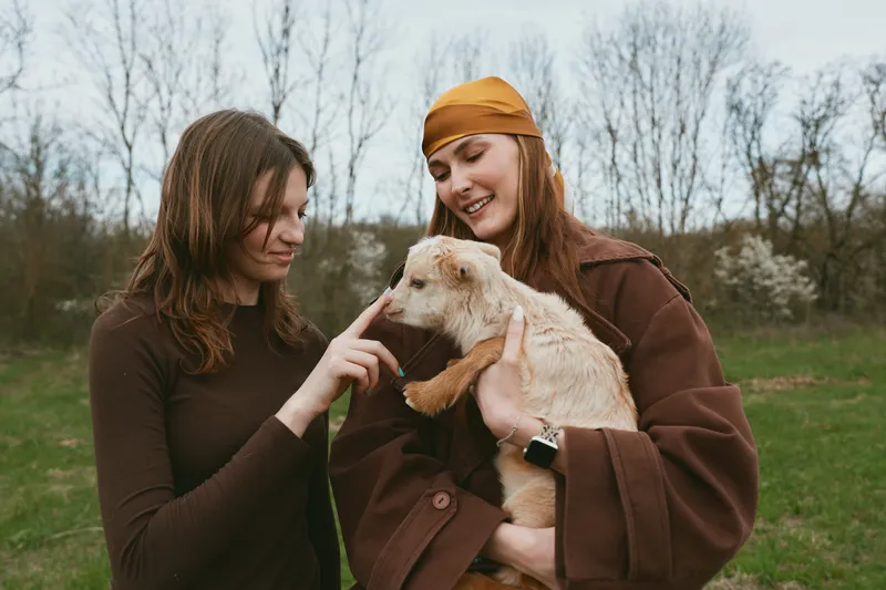 Two women outdoors with a goat kid, sharing a joyful moment, candid lifestyle scene, friendly friendship, laughter, gentle touch, natural light, earthy colors, grassy field, playful interaction