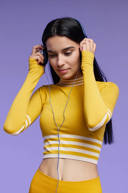 young woman sport fitness dressed in yellow activewear adjusting earphones before workout session with focused expression on purple background