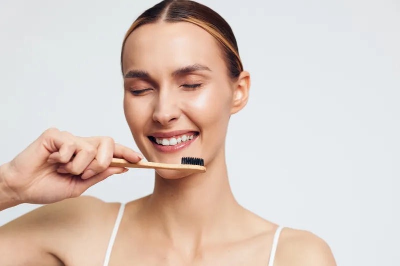 Young woman with closed eyes smiling gently while holding a bamboo toothbrush near her face, natural skincare routine, fresh clean look, white background, personal hygiene concept