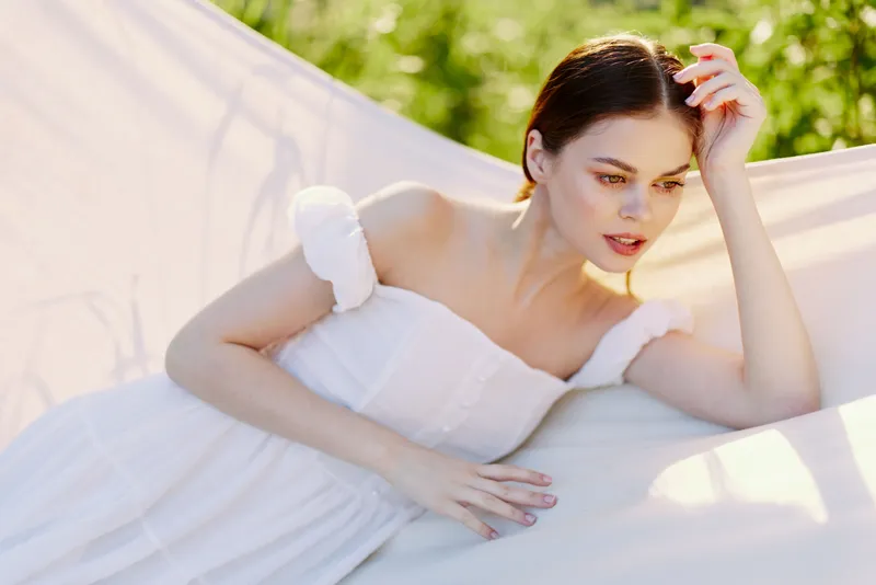 Young woman in a white dress lounging outdoors, displaying serenity and beauty in a sunlit green environment, evoking summer vibes with a soft palette