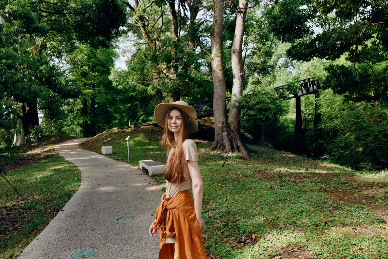 Woman walking in park on gravel path among tall trees, wearing a wide hat and orange skirt, smiling at camera. Outdoor nature portrait, summer leisure and relaxed lifestyle scene.